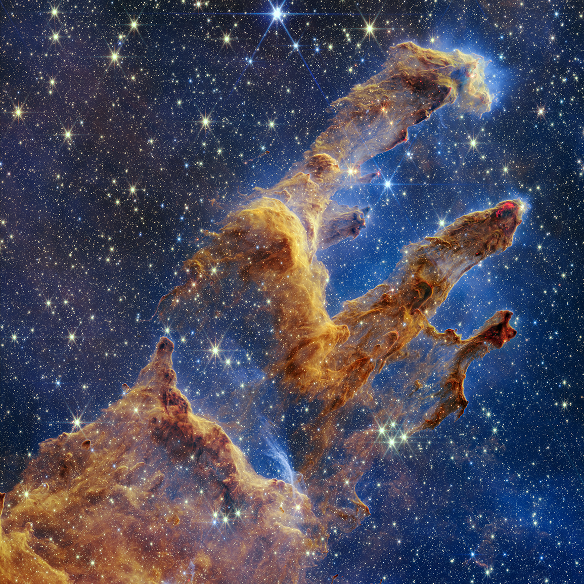 The Pillars of Creation in infrared light. The dust looks thinner and glows gold, and many more stars are visible shining through it.